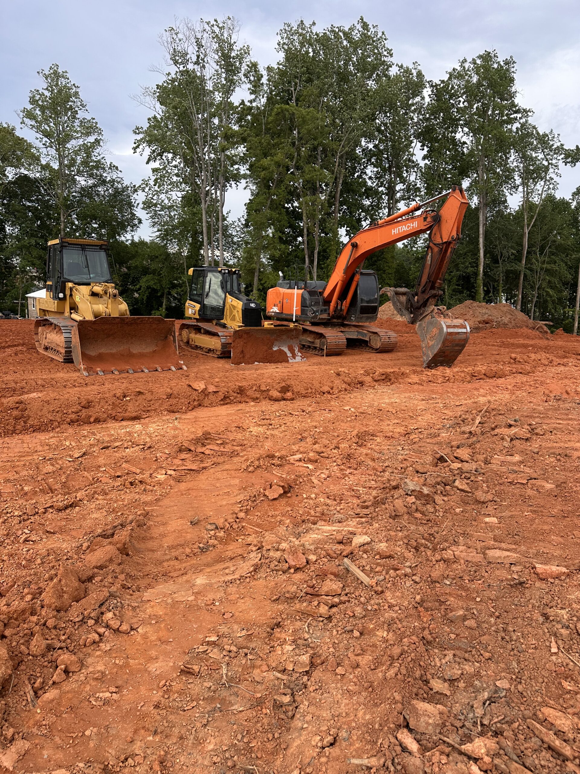 Bulldozers and an excavator are parked on a large area of freshly excavated red soil, with trees visible in the background.