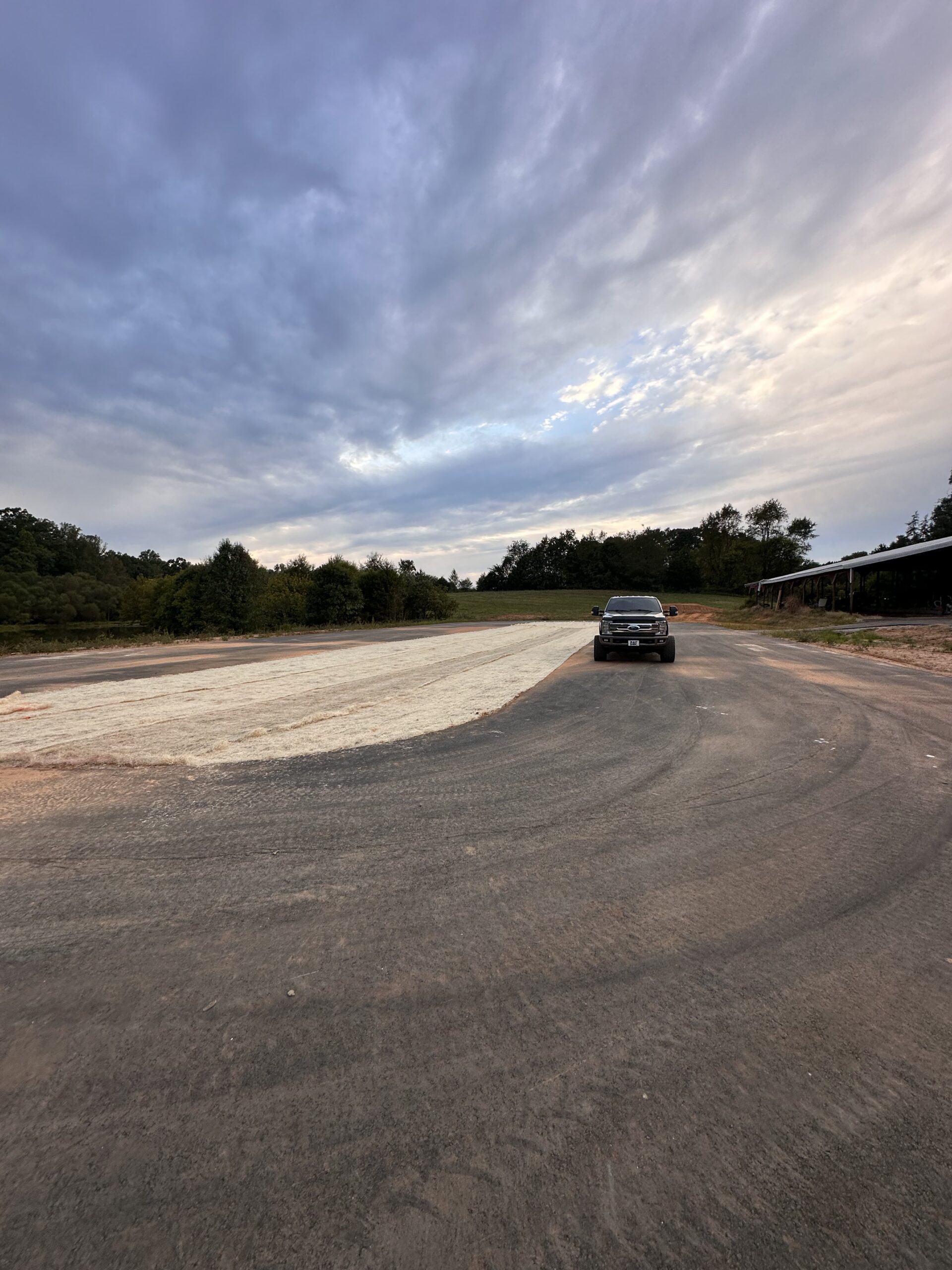 A black pickup truck is parked on a large, mostly empty lot with a mix of paved and gravel surfaces under a cloudy sky. Trees and a shed are visible in the background.