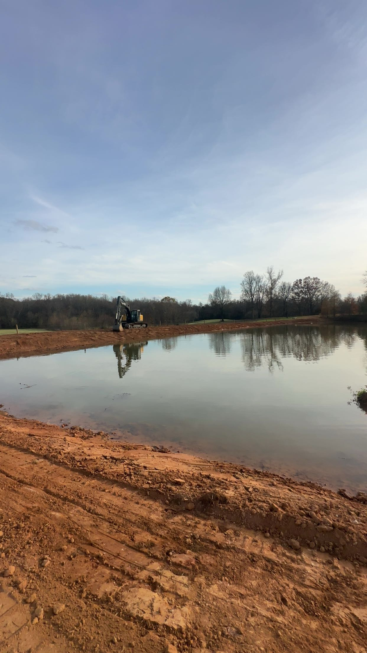 A small excavator is parked near the edge of a calm, reflective pond, with bare trees and a dirt embankment in the background under a mostly clear sky.